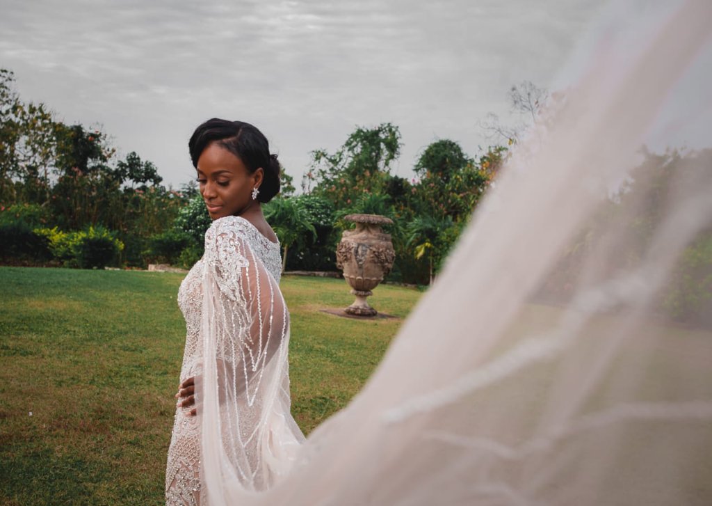 Bride in beaded cape gown, eyes downcast, veil billowing in foreground, estate gardens and stone urn behind
