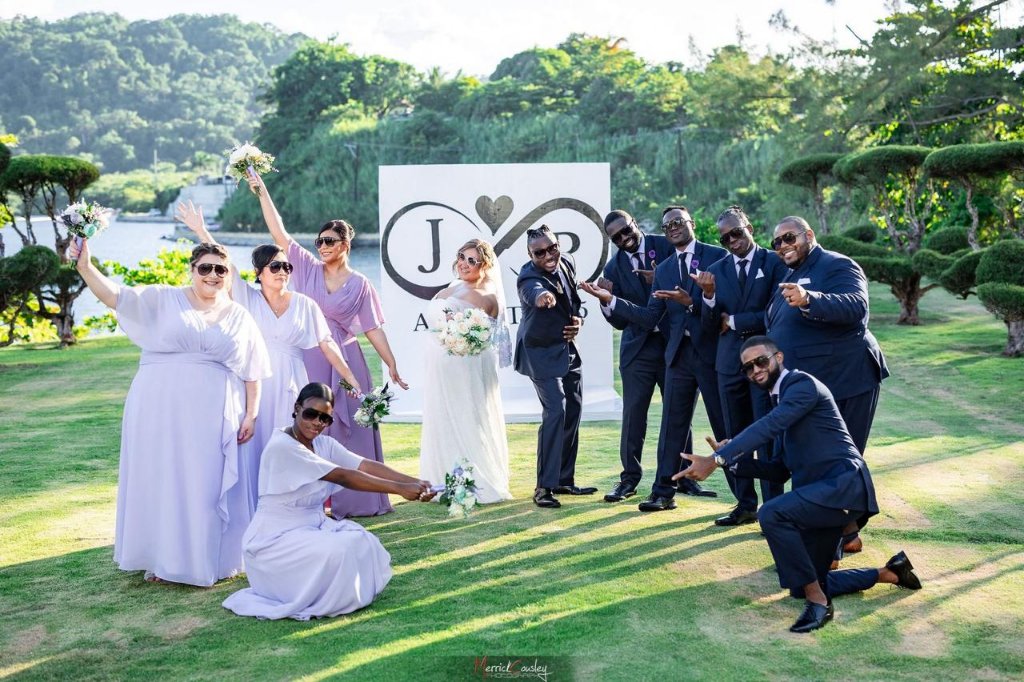 Full wedding party celebrating in front of J&P monogram sign, bridesmaids in lavender and groomsmen in navy