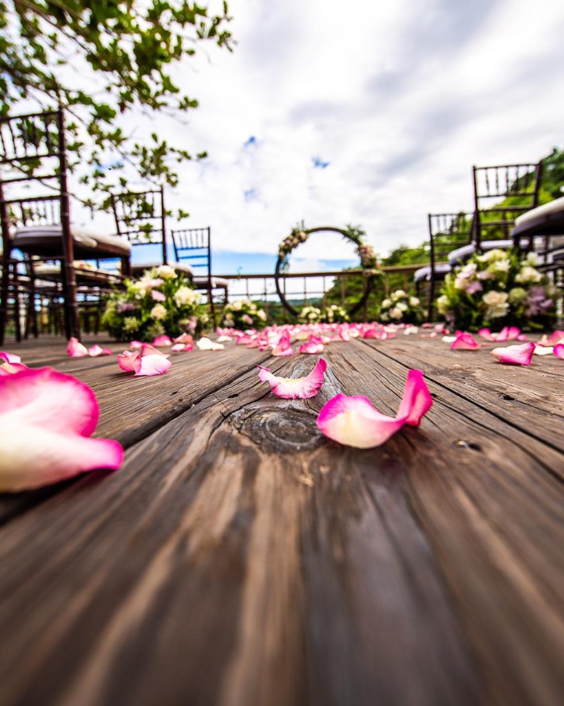 Ground-level shot looking down petal-scattered aisle toward circular arch, pink petals in sharp focus on dark wood deck