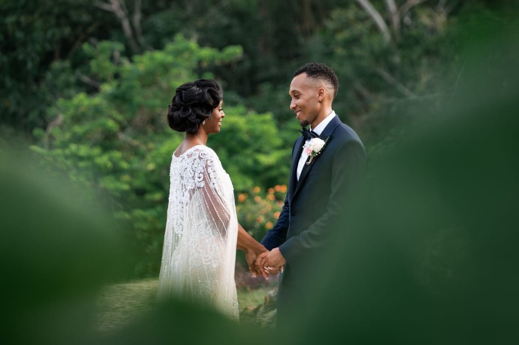 Couple holding hands face-to-face in lush tropical garden, framed by soft green foliage, bride in beaded cape gown, groom in black tuxedo