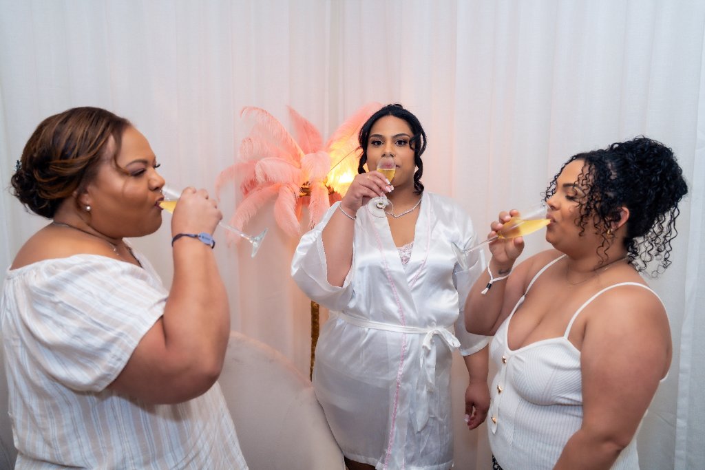 Bride in white satin robe sipping champagne with two bridesmaids in front of pink feather arrangement and white draping
