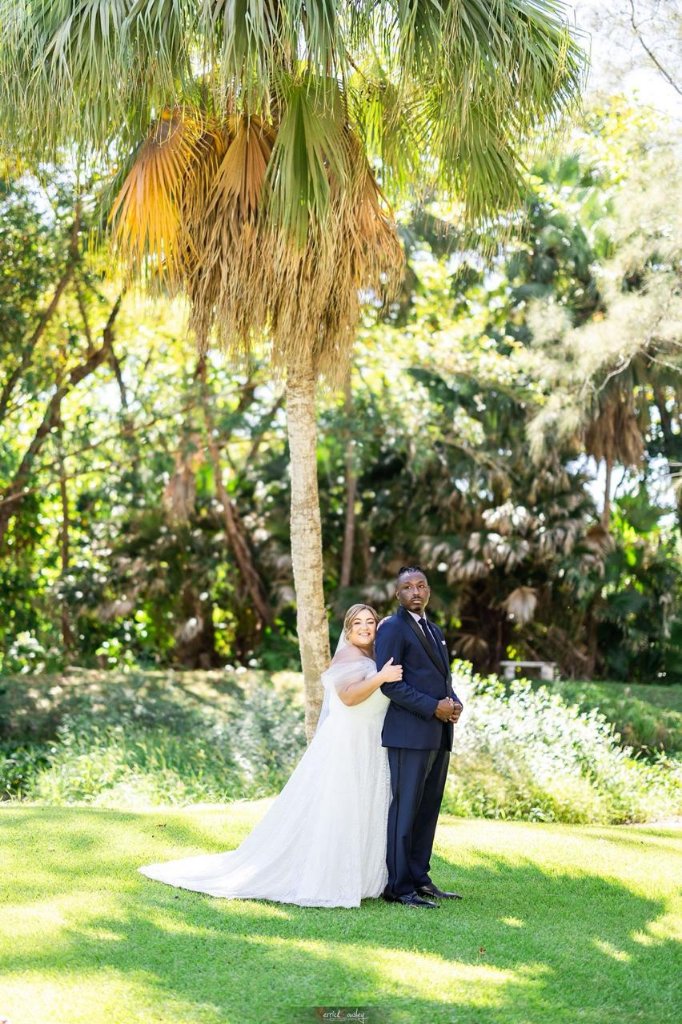 Couple under tall palm tree, bride hugging groom from behind, both facing camera