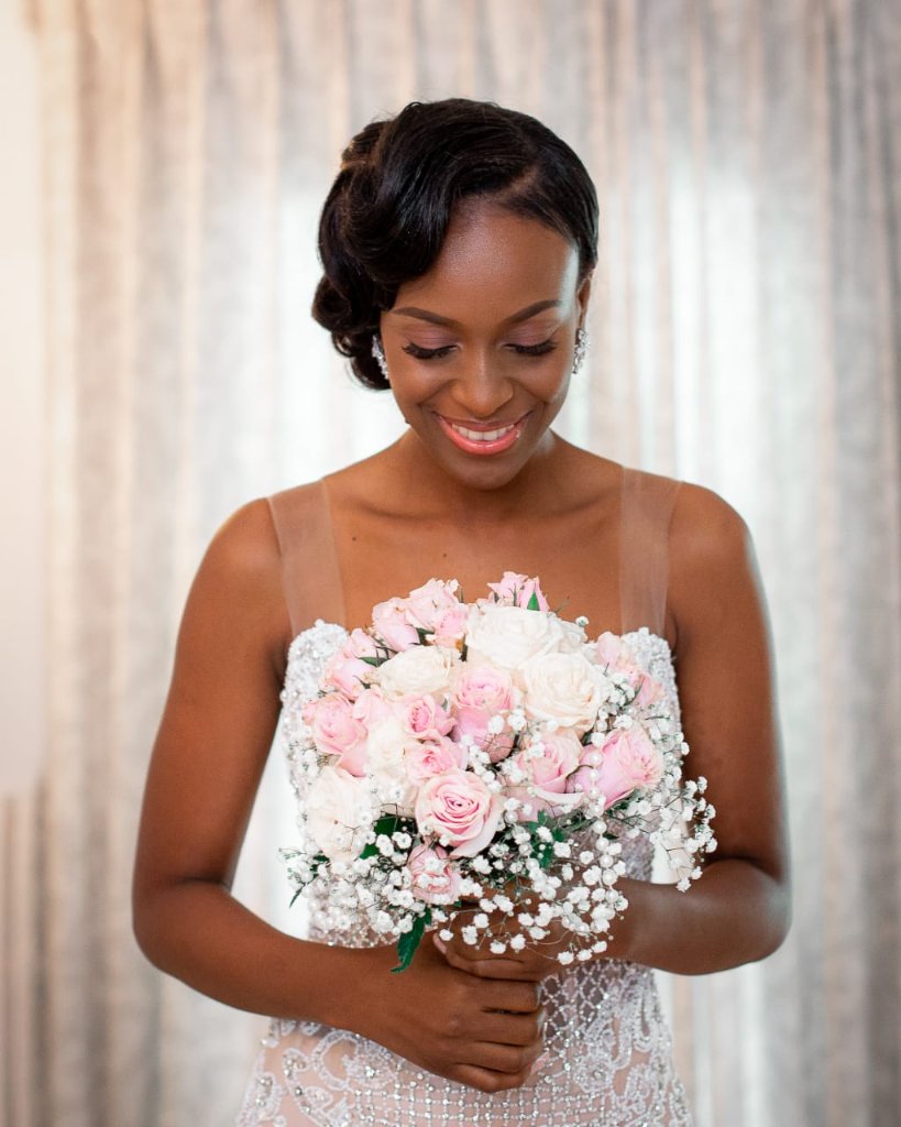 Bride smiling down at her blush bouquet of pink roses and baby's breath, beaded gown detail in full view
