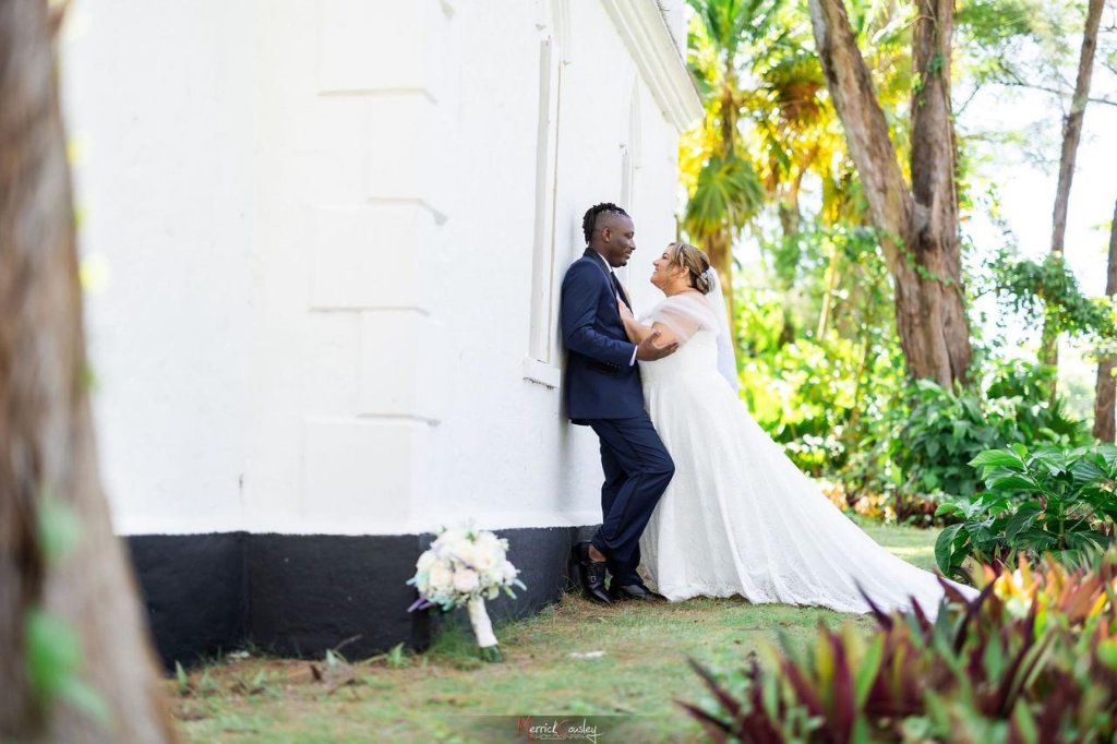 Couple leaning against white chapel wall in tropical garden, gazing at each other
