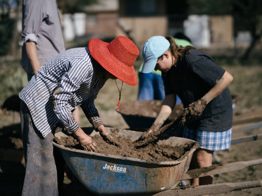 Two volunteers mixing wet adobe through buffelgrass with hands in a wheelbarrow