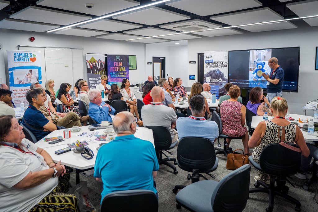 Wide view of the full room with attendees seated at hexagonal tables during a presentation