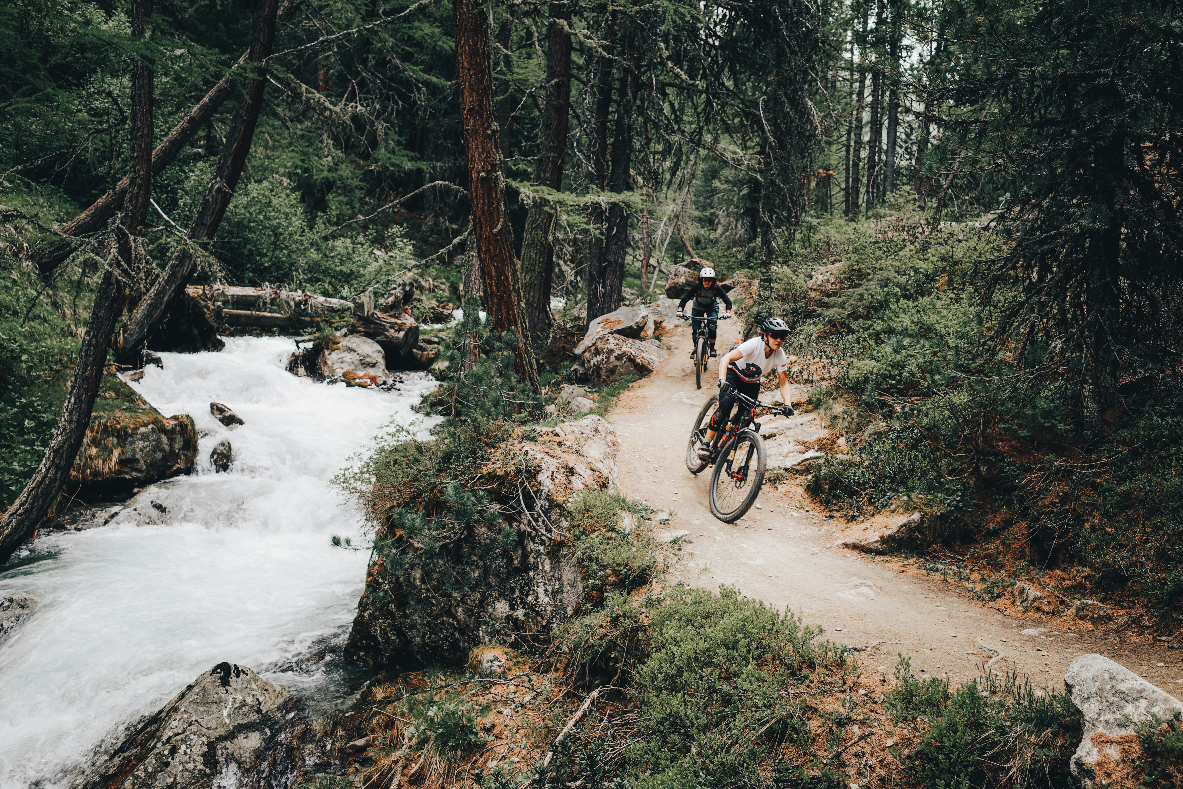 Two mountain bikers riding a single-track trail beside a rushing mountain stream