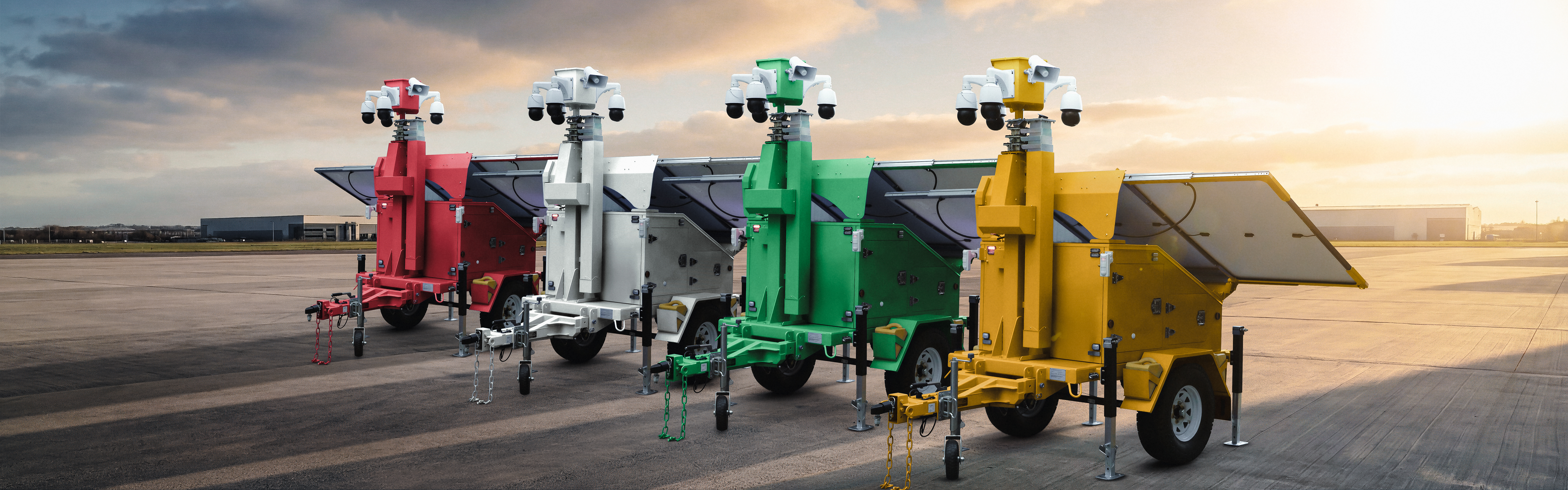 Four mobile surveillance trailers — red, white, green, and yellow — lined up on an industrial lot at sunset