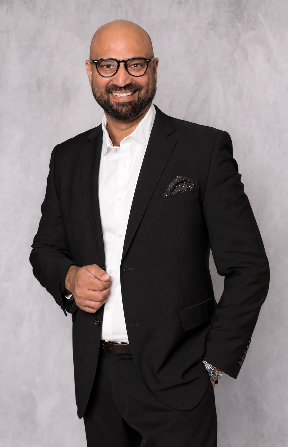 Professional medium-shot portrait of a bald man with beard and dark-rimmed glasses in black suit with white shirt against a mottled grey studio backdrop, warm confident smile