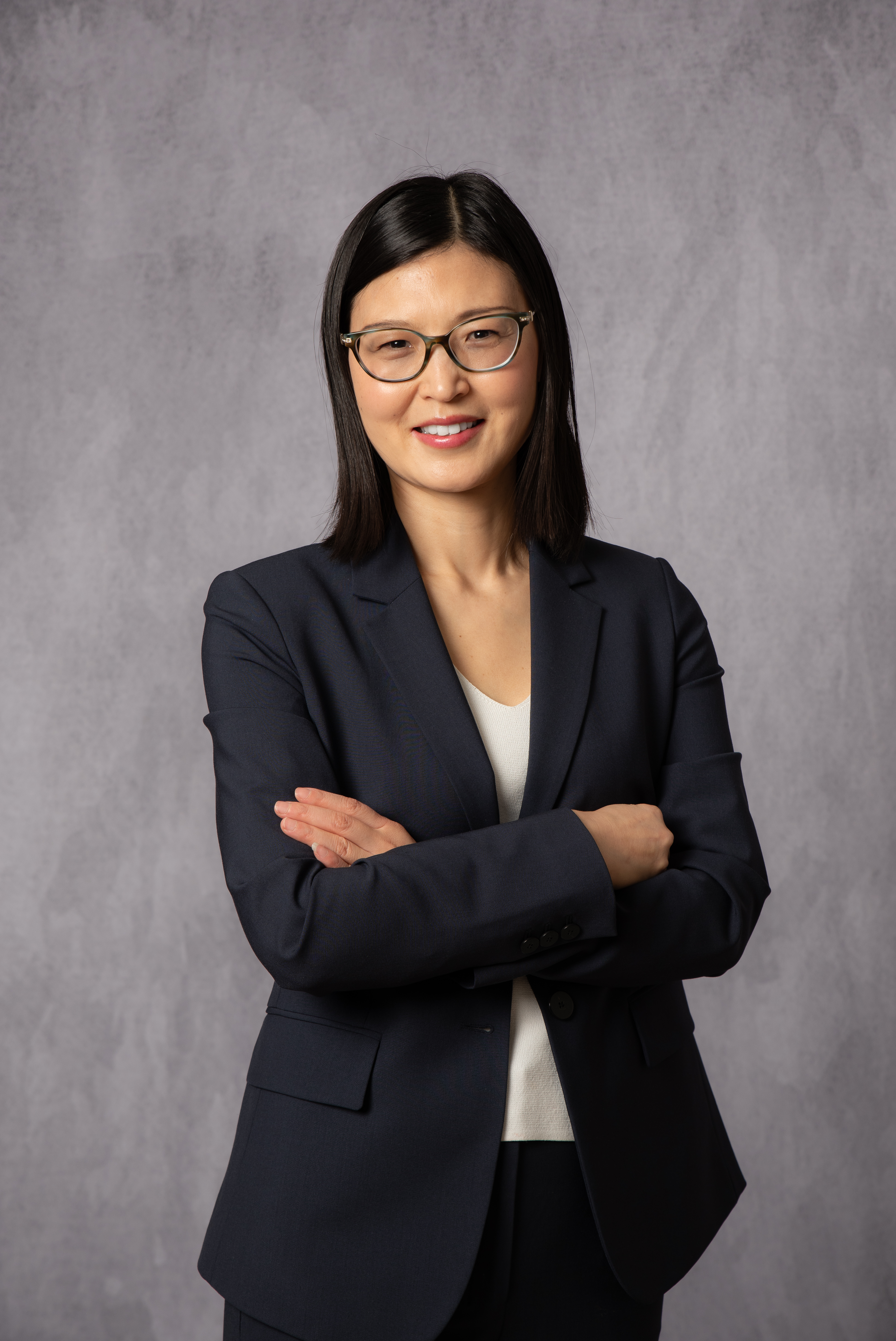 Professional corporate portrait of a woman with dark shoulder-length hair and glasses, navy blazer over white top, arms crossed, standing against a mottled grey studio backdrop