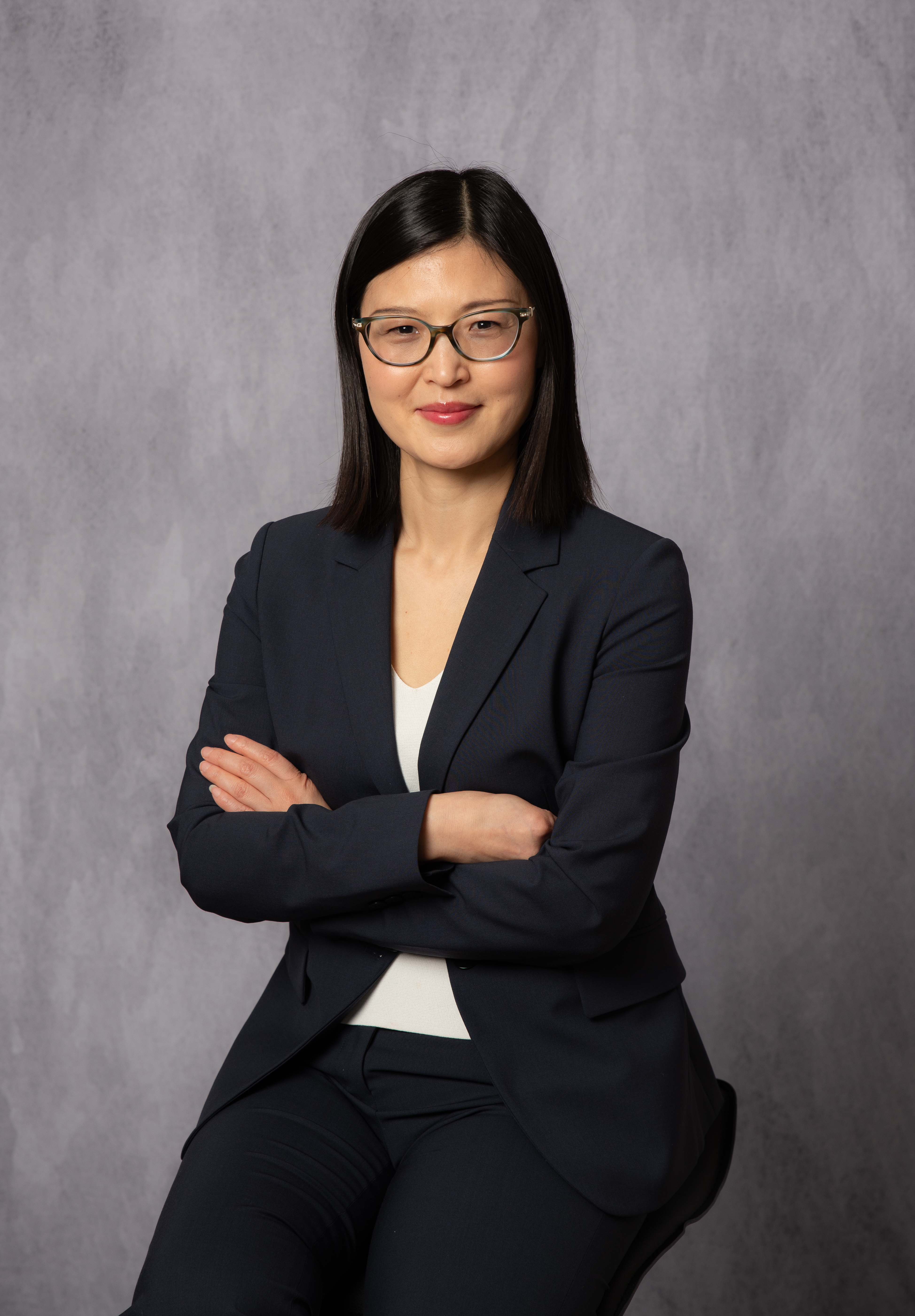 Professional studio headshot of a woman with black shoulder-length hair and glasses, navy blazer over white top, seated with arms crossed against a mottled grey studio backdrop, warm friendly smile