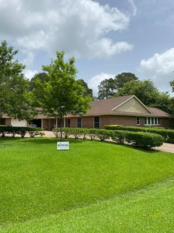 Single-story red brick ranch with dark brown roof and Roofing Solutions yard sign