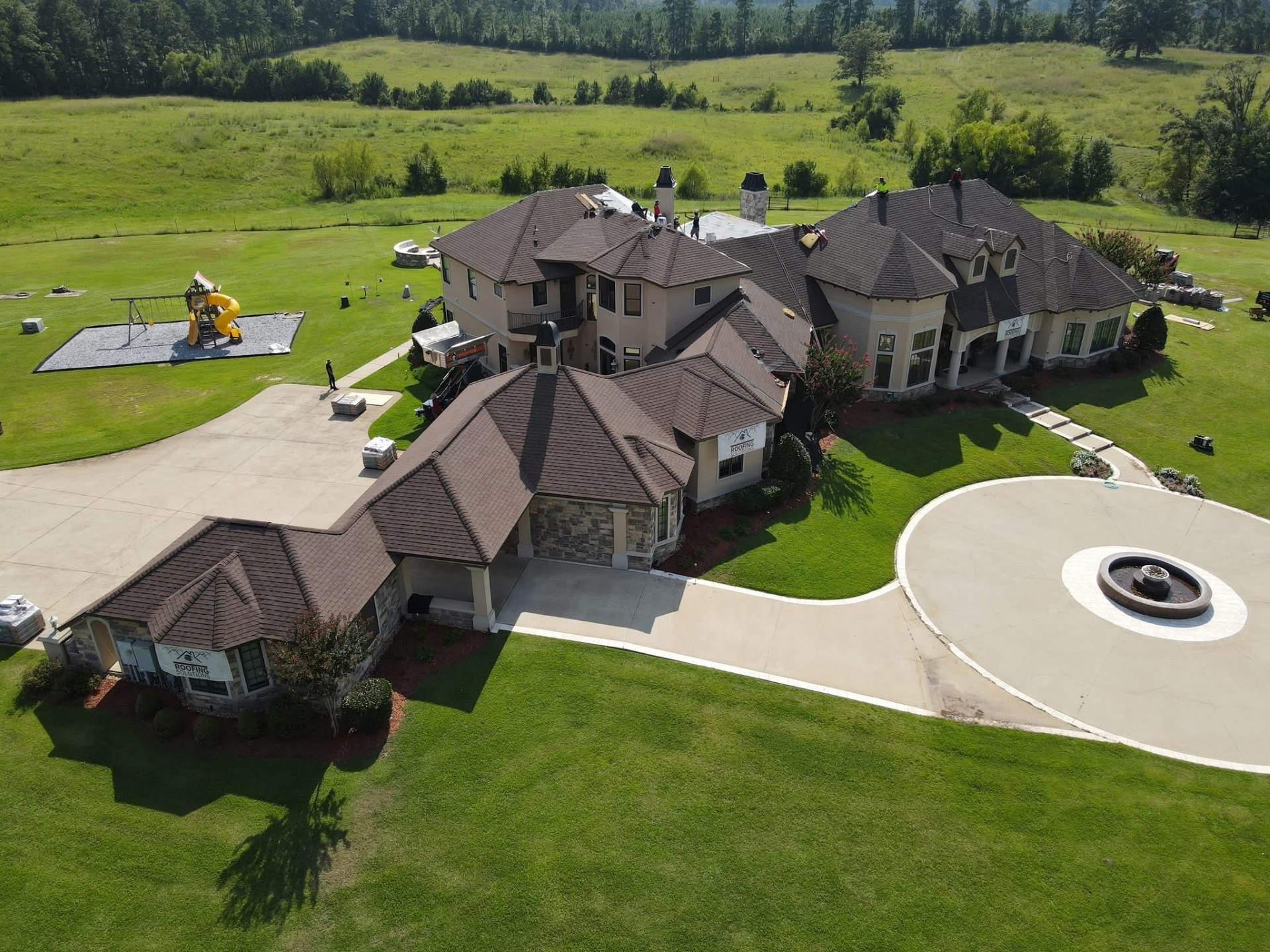 Aerial of multi-gabled estate with crew on the roof and Roofing Solutions banners