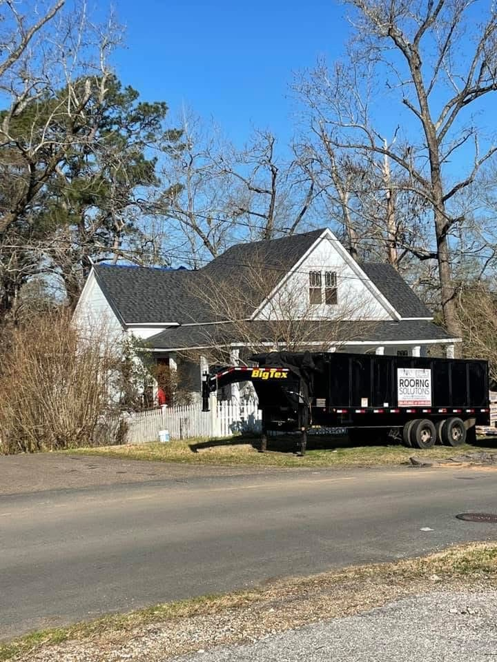 White single-story home with blue tarp on the eave and dump trailer staged