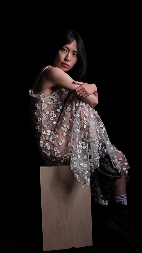 Portrait seated on wooden block in floral lace dress against dark background