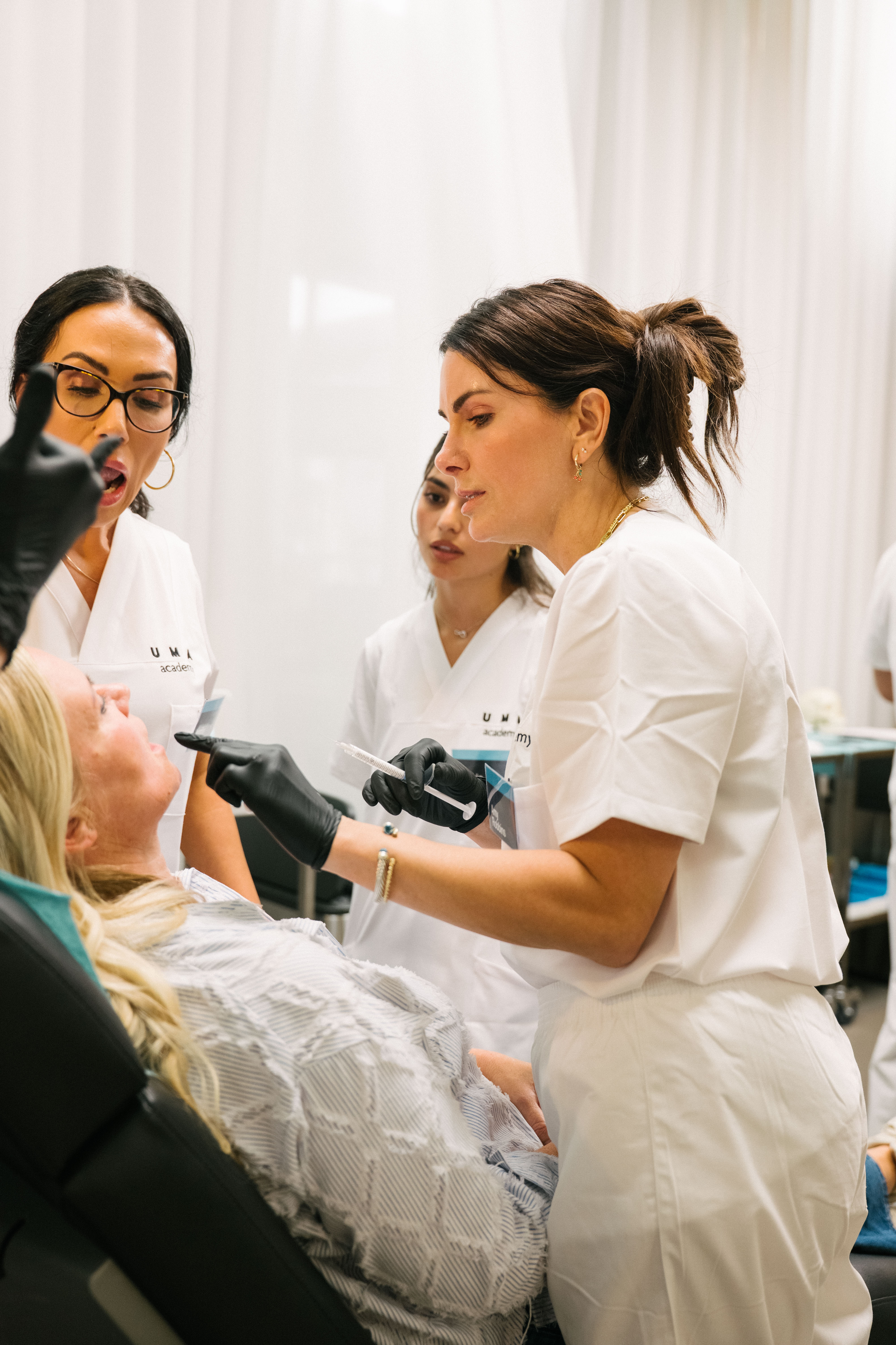 Gloved injector in white scrubs preparing to treat a reclined client's lower face during a clinical session