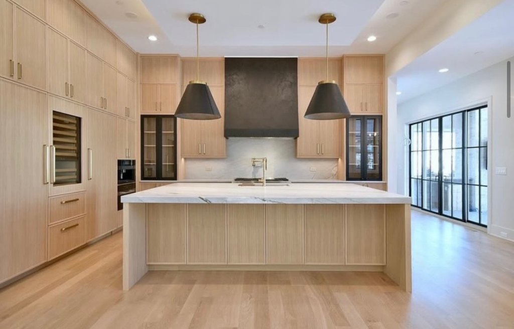 Custom kitchen remodel with light oak flat-panel cabinets, large marble island, black range hood, gold hardware and pendant lights in Dallas TX