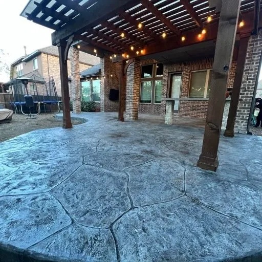 An expansive stamped concrete patio with an irregular stone pattern, featuring a wooden pergola adorned with string lights, creating an inviting outdoor living space in DFW.