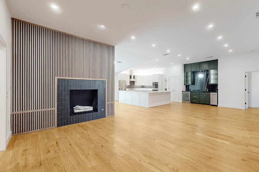 Spacious open concept living room featuring newly installed light oak hardwood flooring, contemporary fireplace with black tile surround, slatted wooden accent wall, and modern kitchen by 448 Developments Dallas TX