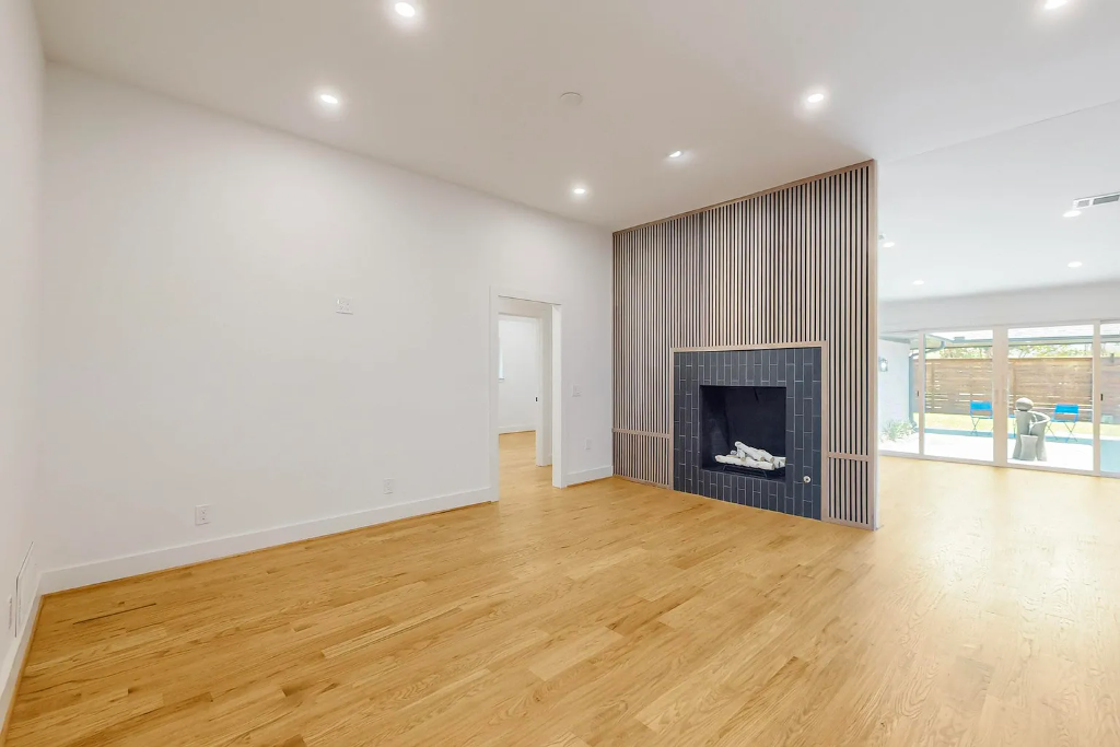 Newly installed light hardwood flooring in a bright open living room with a modern tiled fireplace, slatted accent wall, and glass sliding doors to backyard by 448 Developments Dallas TX