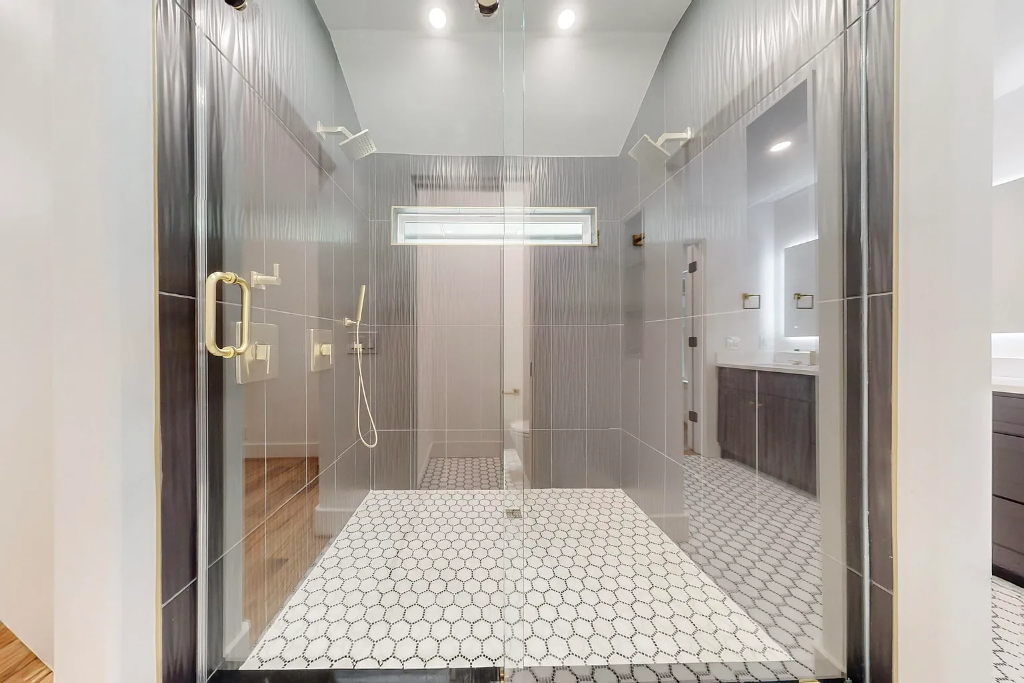 Large luxury walk-in shower with grey vertical tile walls, hexagon tile floor, glass enclosure and gold fixtures in DFW
