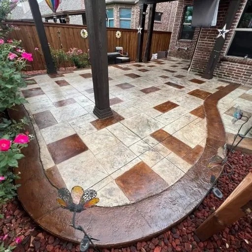 A decorative two-tone stamped concrete patio featuring square patterns and a curved border, surrounded by lush landscaping, ideal for custom patio projects across DFW.