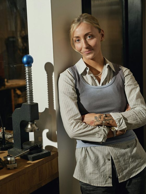 A small business owner stands with her arms crossed in her workshop, beside a bench press and tools.