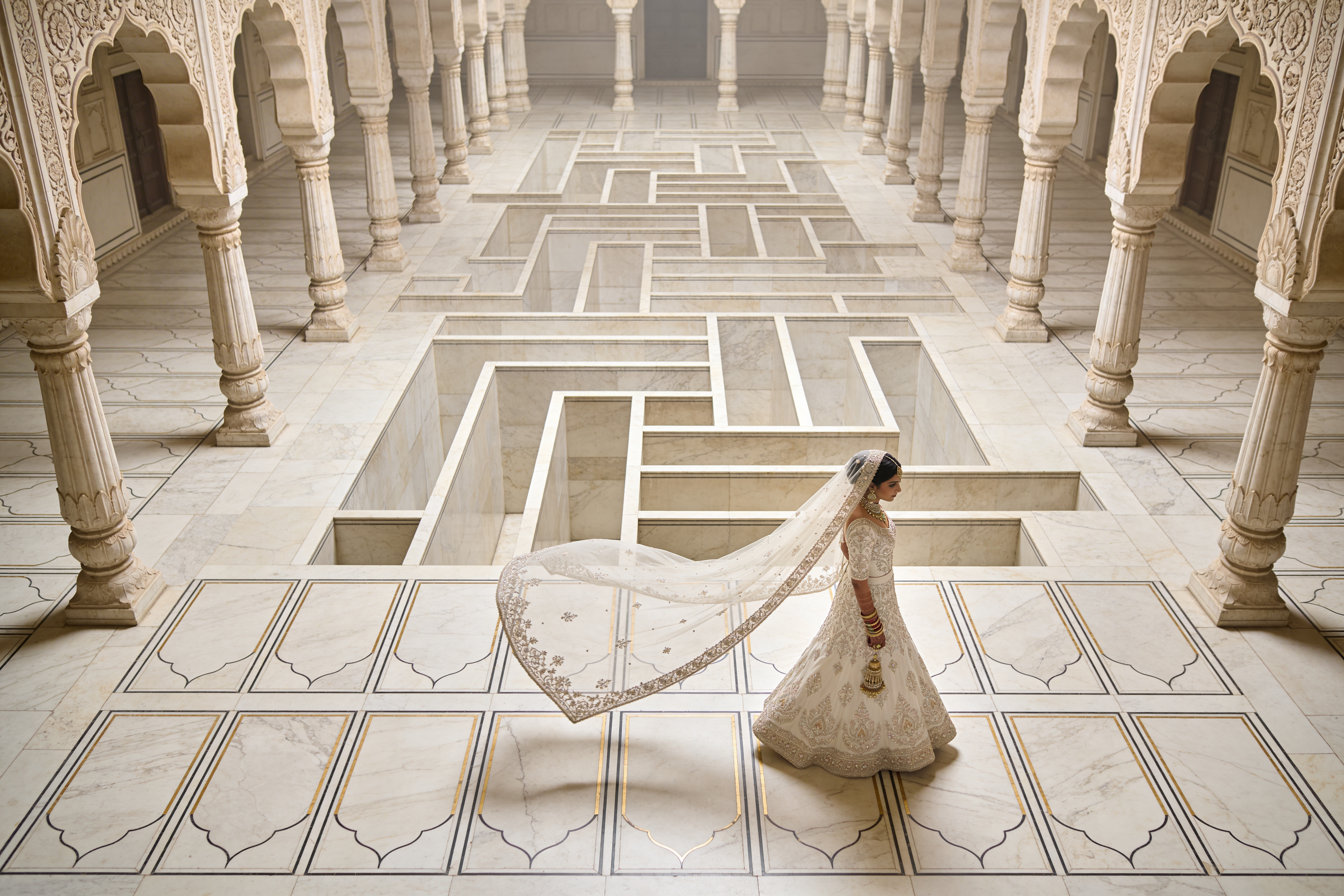 Bride wearing an embroidered ivory lehenga and long embroidered veil walking across a carved marble palace courtyard with colonnades and geometric recessed floor patterns, photographed from above.