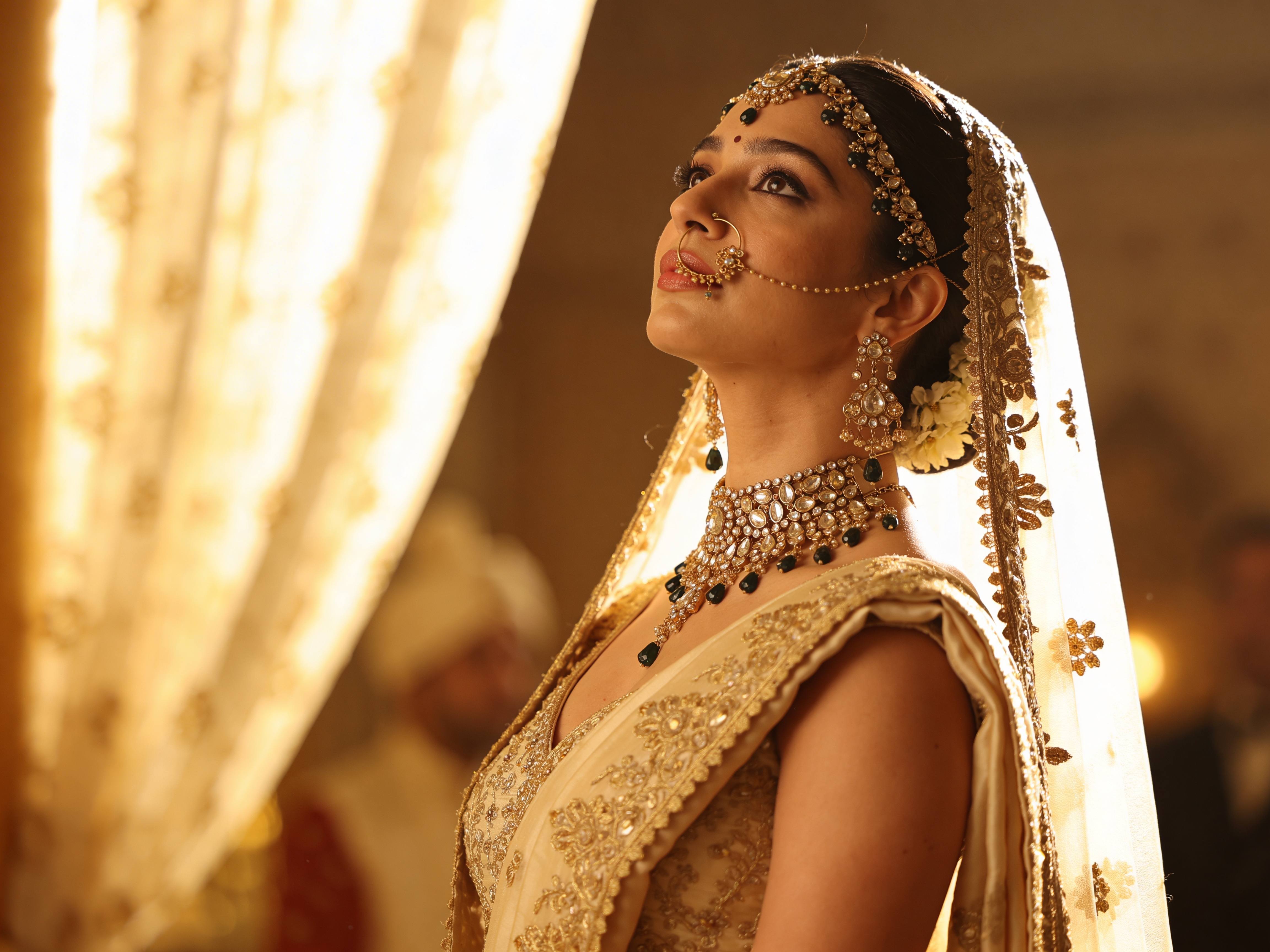 Bridal portrait of a South Asian bride in a gold-embroidered lehenga and sheer dupatta, wearing ornate Kundan and emerald jewelry including a choker, jhumkas, matha patti and nath, lit by warm backlight in a heritage interior.