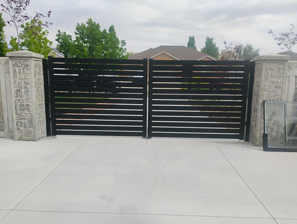 Black horizontal-slat double-swing gate mounted between grey stacked-stone pillars on a concrete driveway