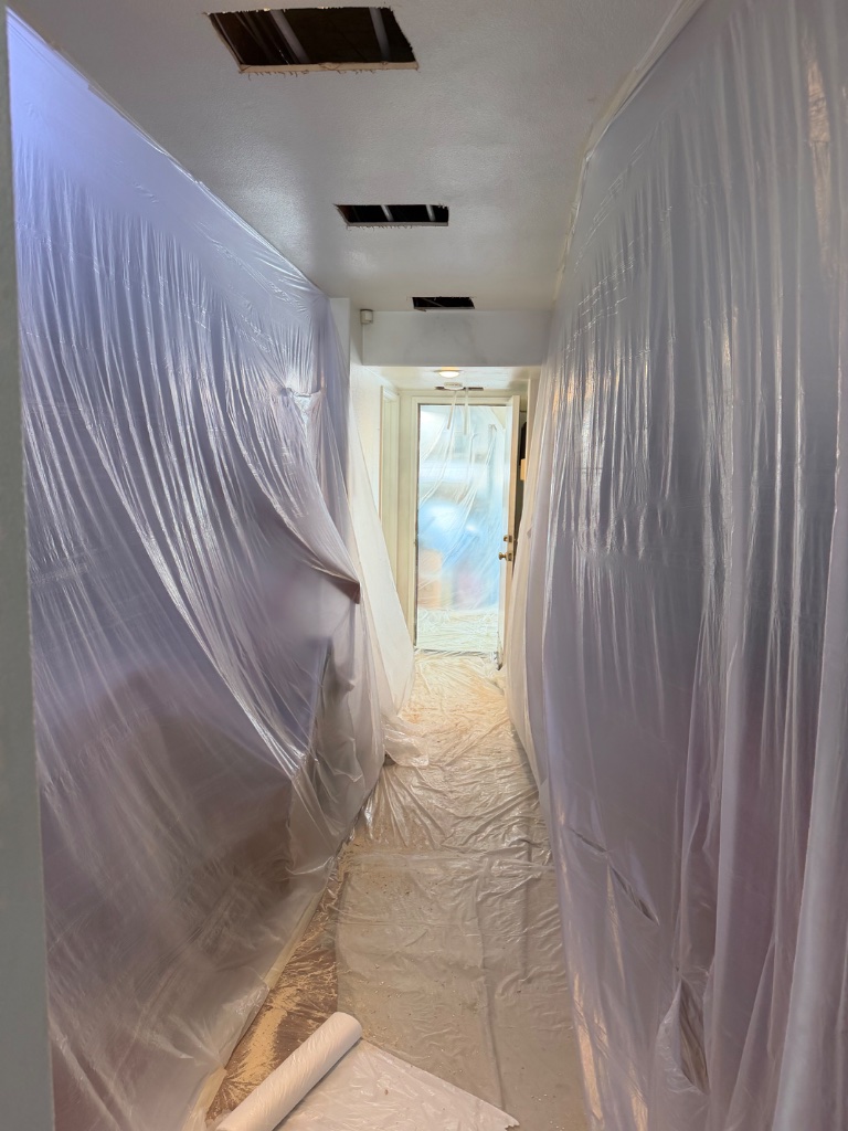 Hallway fully contained with plastic sheeting — ceiling access holes cut for pipe routing