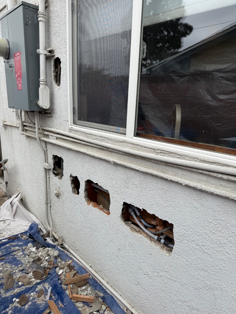 Exterior stucco wall with multiple access holes and new pipe lines visible, electrical panel above