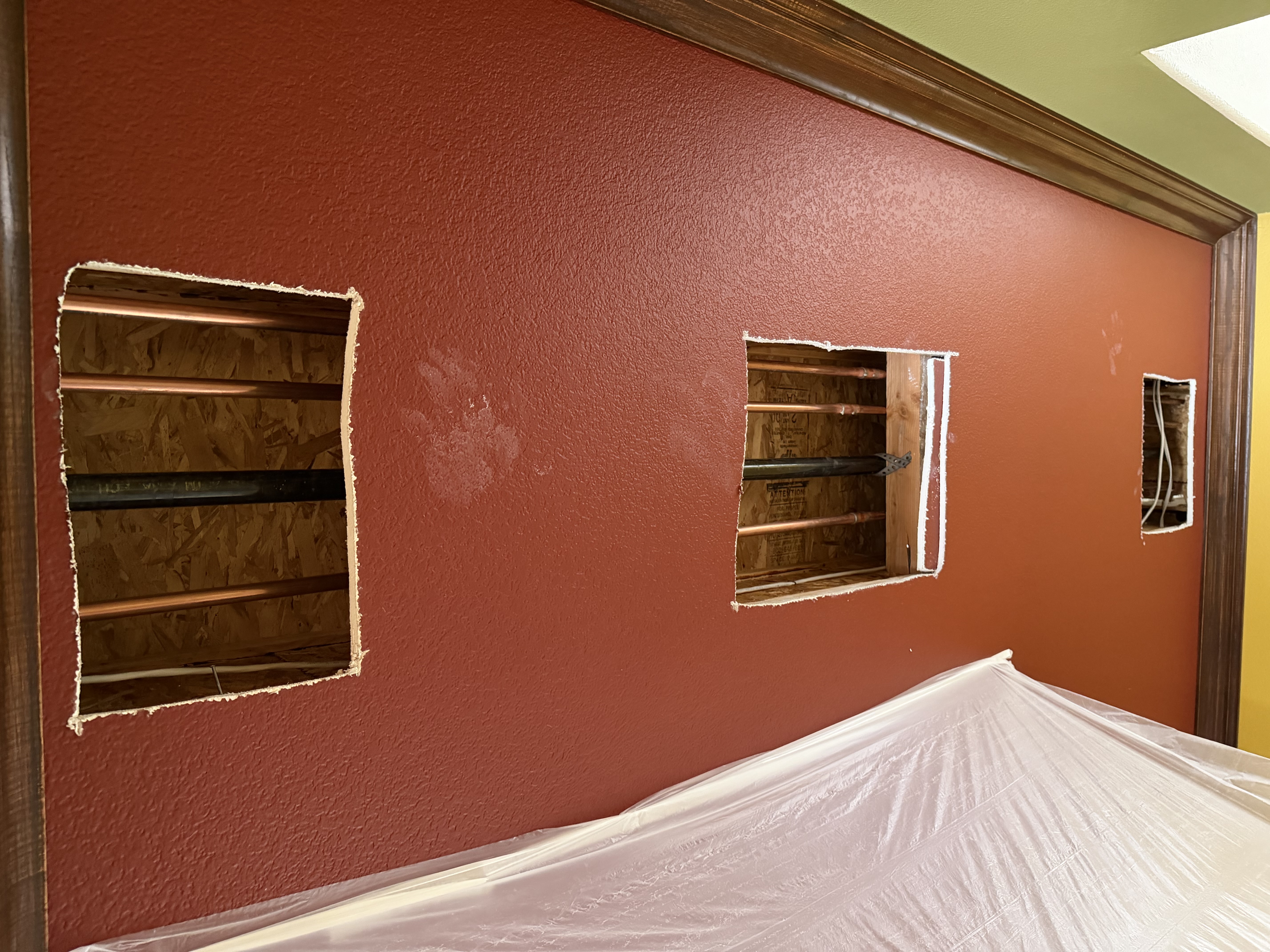 Completed copper pipe installation in interior wall with access cutouts and surface protection