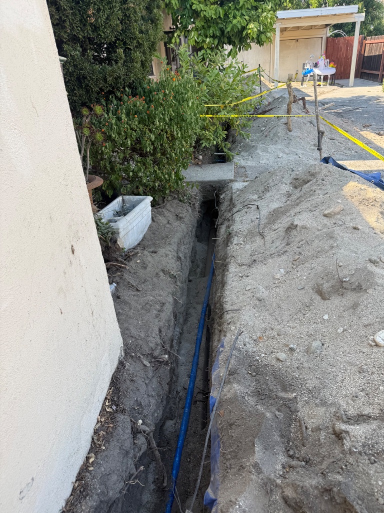 Deep mainline trench alongside home — new blue PEX replacement line laid in