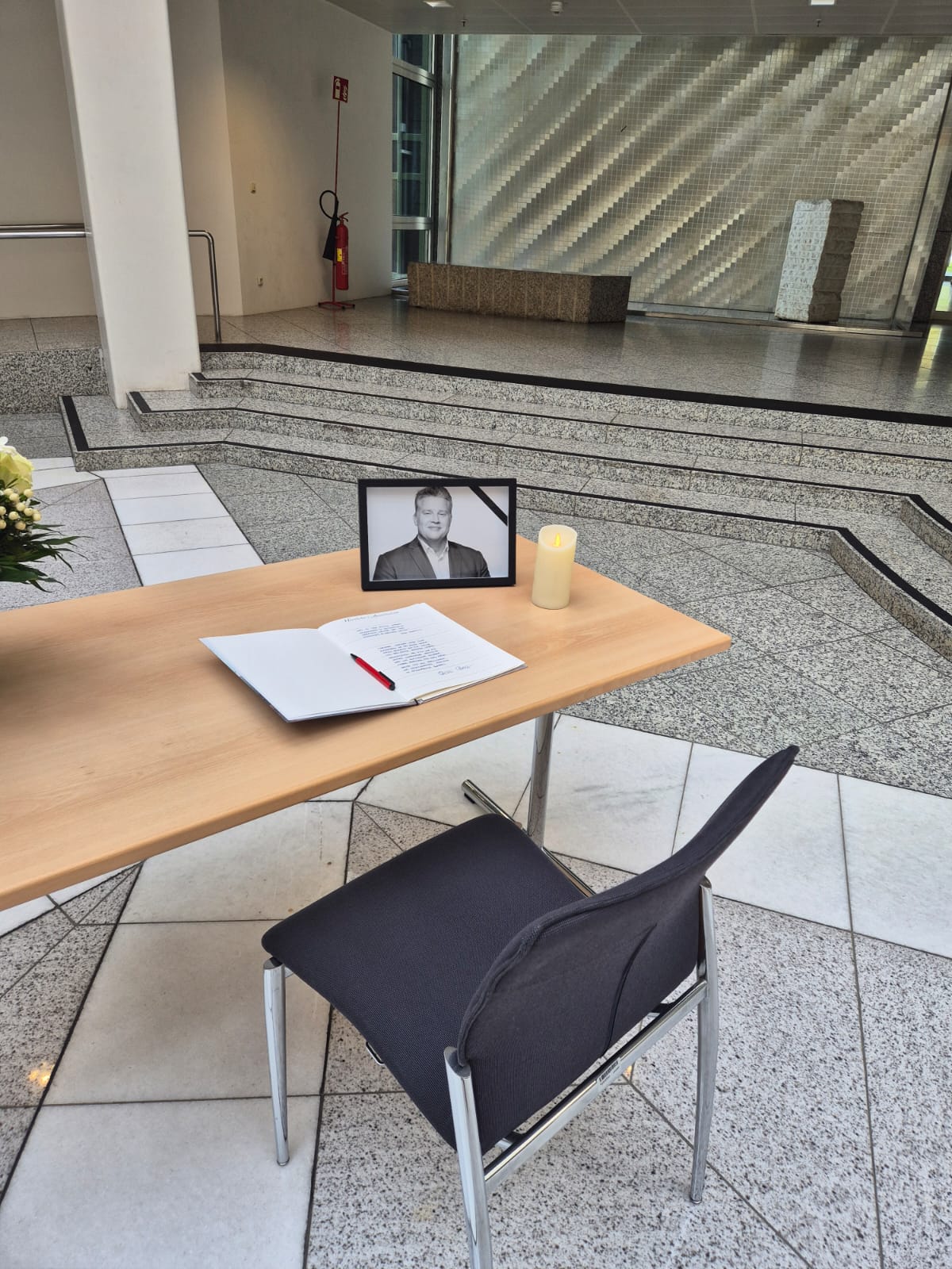 Auf einem Tisch liegen ein Kondolenzbuch und ein roter Stift. Daneben stehen ein gerahmtes Porträt von Carsten Träger und eine brennende Kerze.
