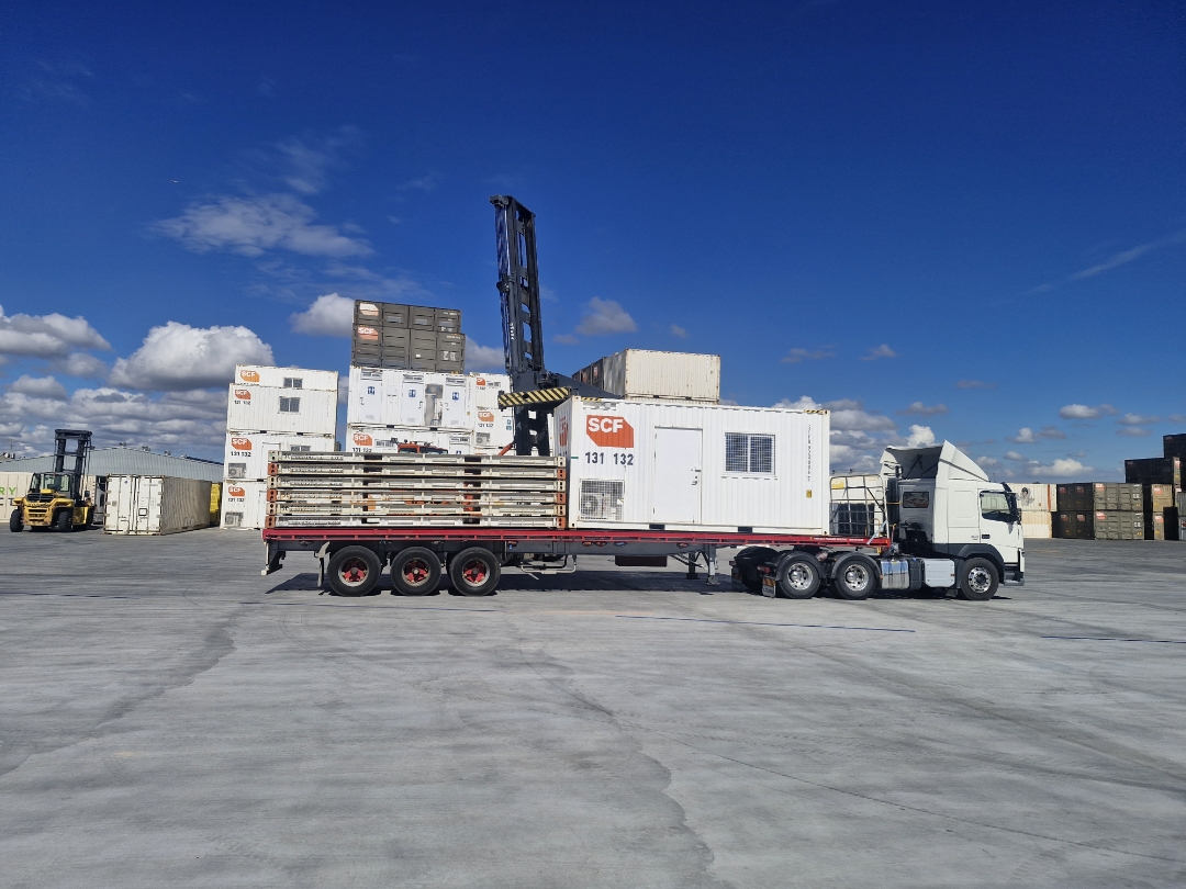 White AFM prime mover hauling an SCF modular unit through a container yard with a reach stacker