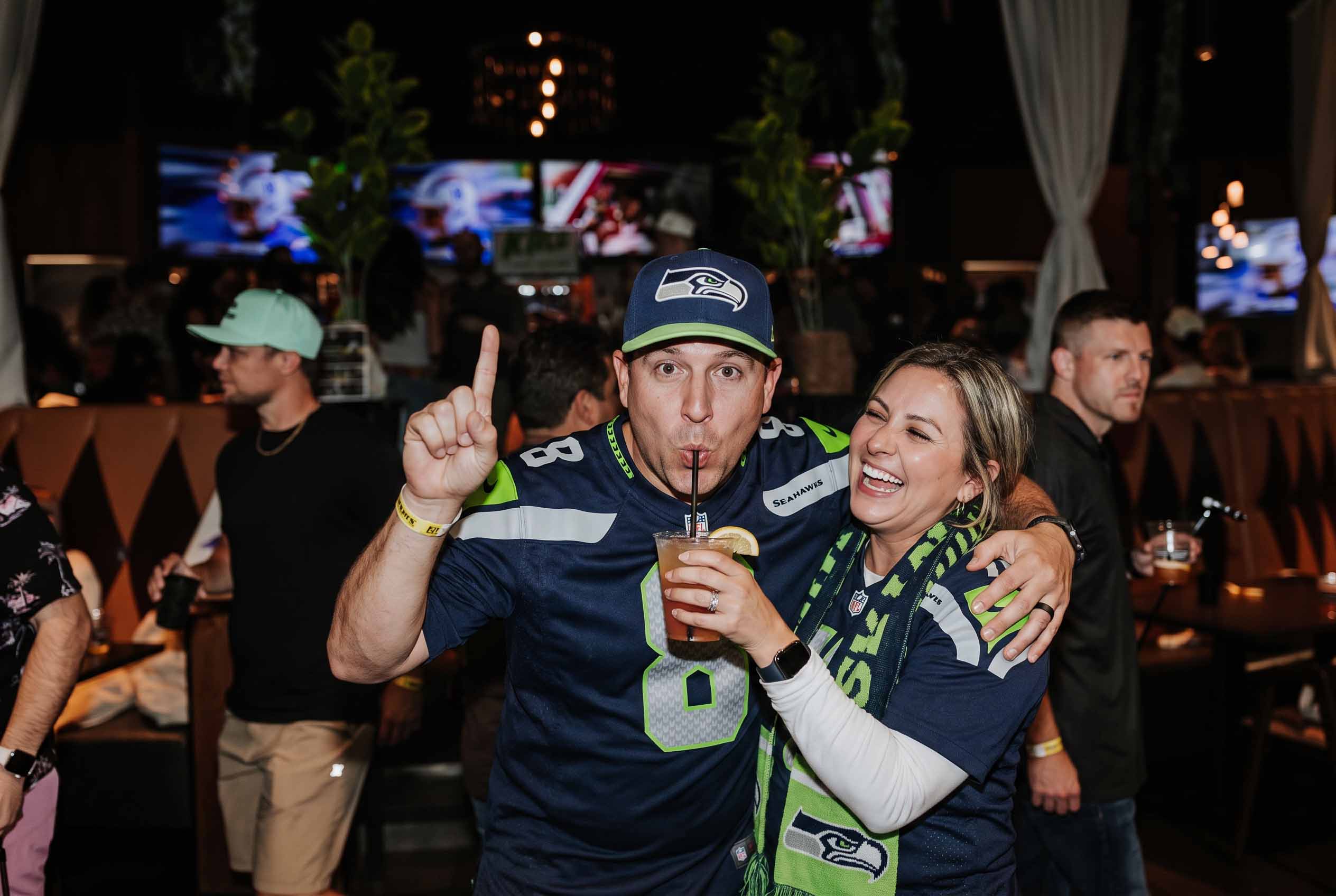 A couple in Seattle Seahawks jerseys smiling and toasting beverages at an indoor putting watch party at Putting World in Scottsdale
