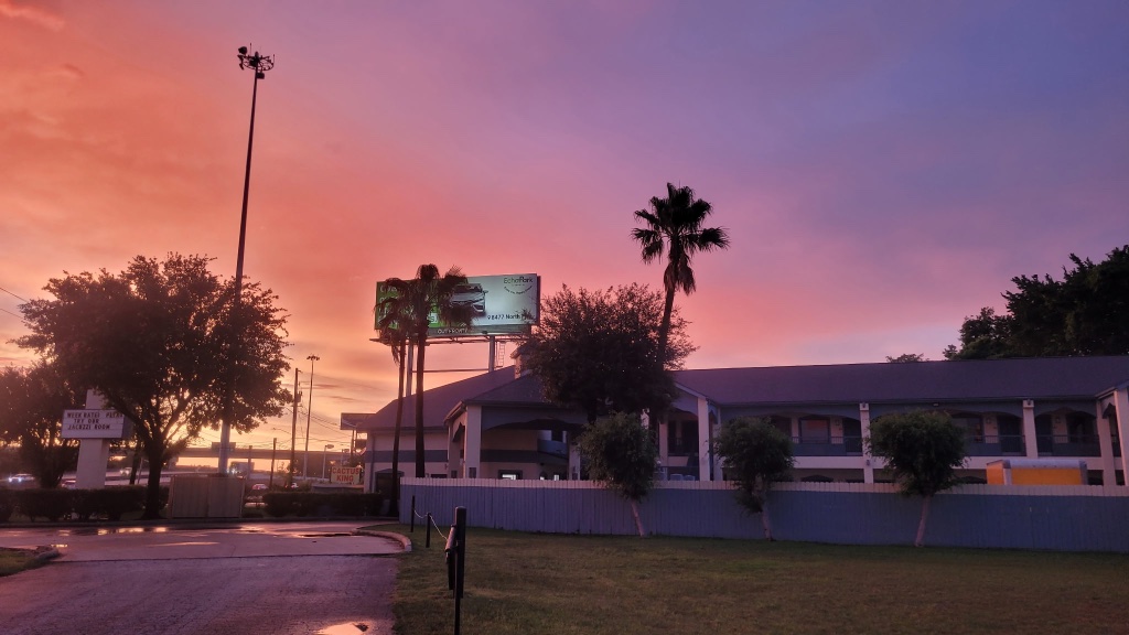 A low-rise building with a gray roof and white walls, surrounded by a gray fence, trees, and a billboard, set against a vibrant sunset sky.