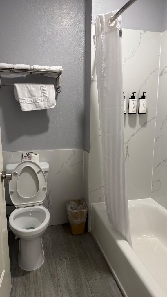 A standard hotel bathroom with a toilet, bathtub with shower curtain, towels, and trash can.