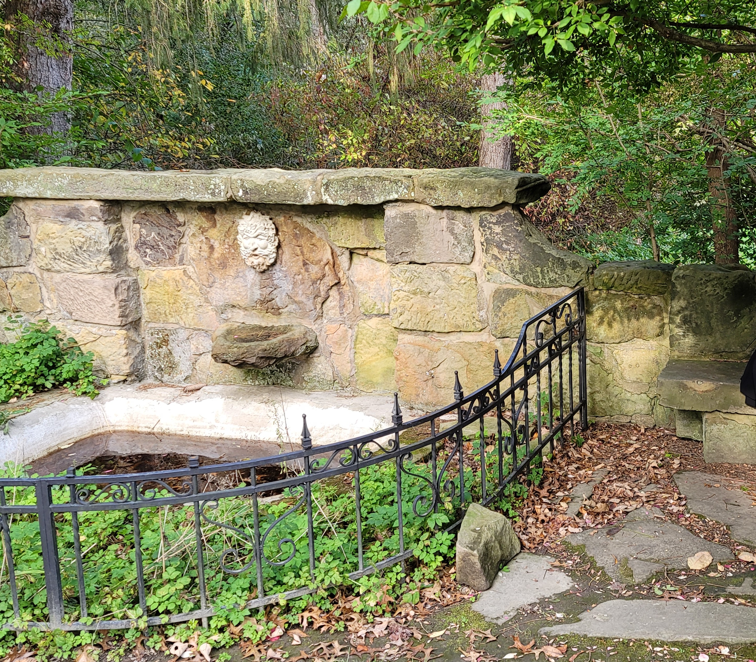 A rustic stone fountain with a carved green-man face and wrought-iron fence in the Quail Hollow gardens
