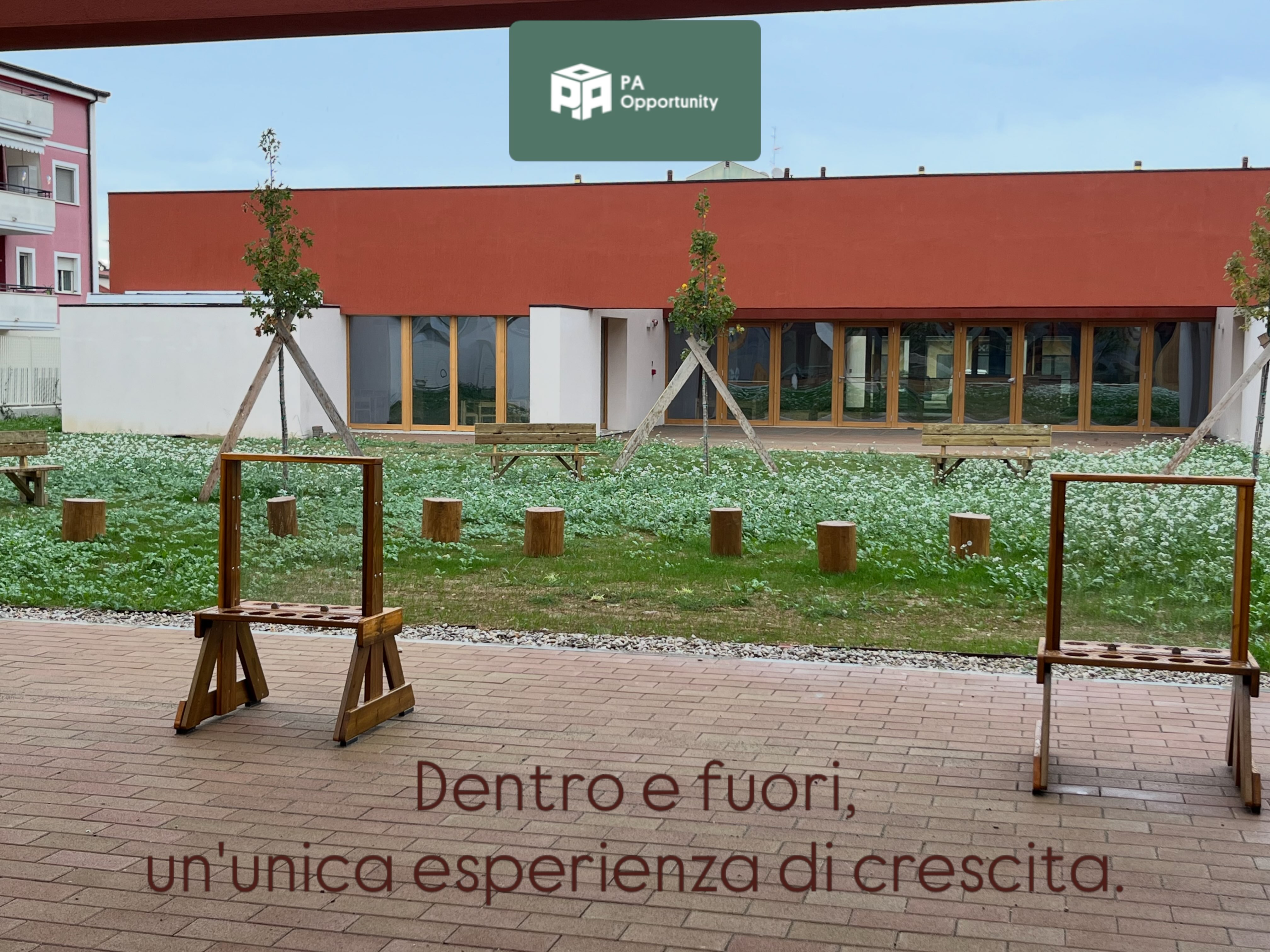 Giardino esterno di un nuovo asilo nido a Montesilvano (PE) arredato con sedute in legno, tronchi, cavalletti e spazi verdi per attività educative all'aperto, con edificio scolastico sullo sfondo.