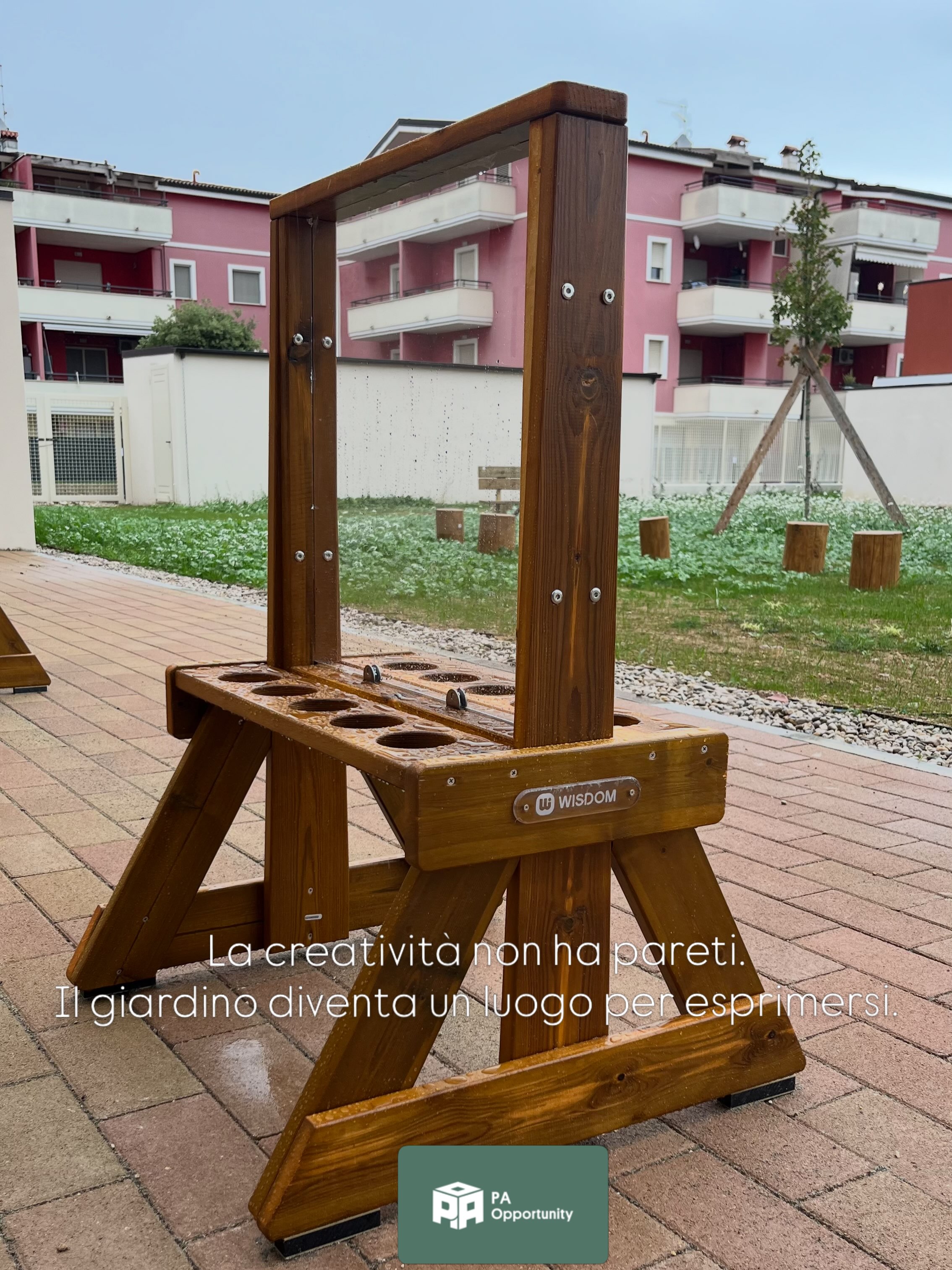 Giardino esterno di un nuovo asilo nido a Montesilvano (PE) arredato con sedute in legno, tronchi, cavalletti e spazi verdi per attività educative all'aperto, con edificio scolastico sullo sfondo.