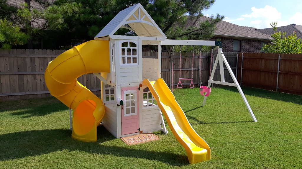 Backyard playset with yellow tube slide and wave slide, pink swings, and playhouse door assembled by Redhawk Handyman Services in Edmond, OK
