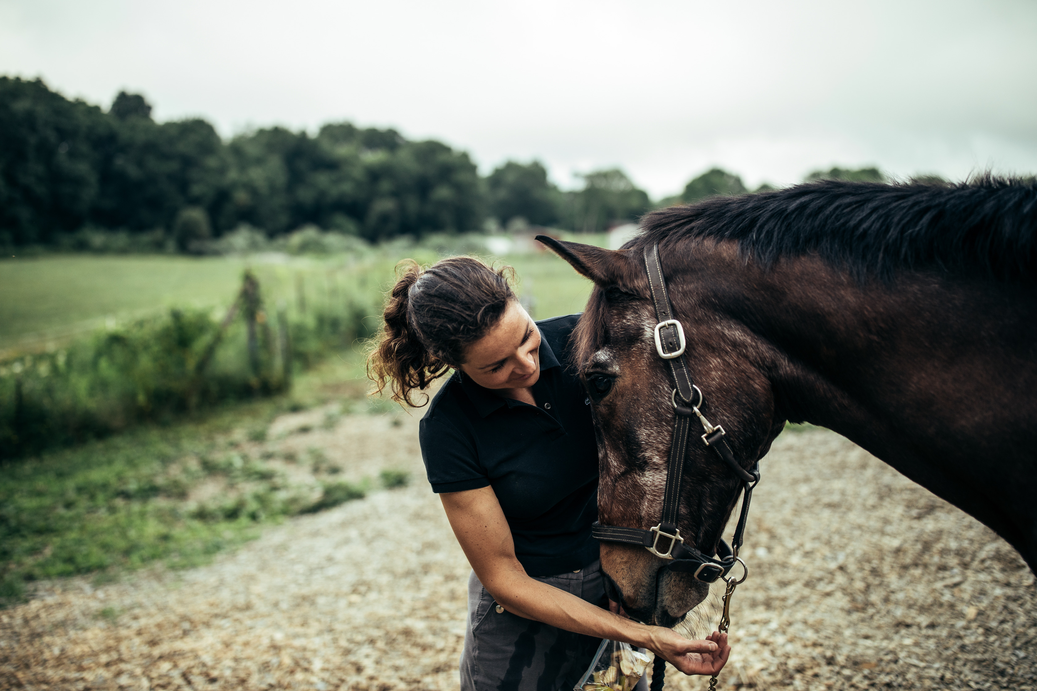 A woman in a dark polo holding a brown horse's lead on a gravel path beside a fenced pasture