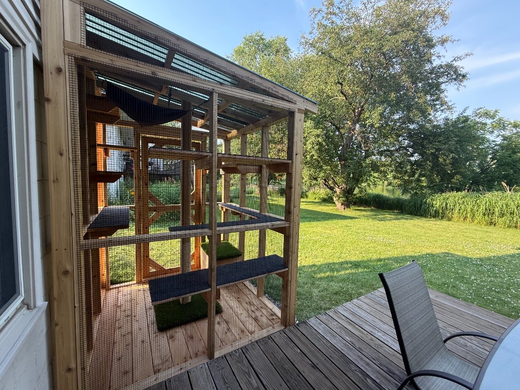 A custom wooden catio on a deck beside a house, surrounded by lush greenery