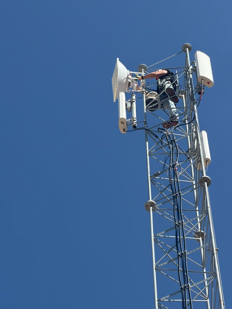 NetWest tower climber servicing a microwave dish at the top of a galvanized steel lattice communication tower under a clear blue sky