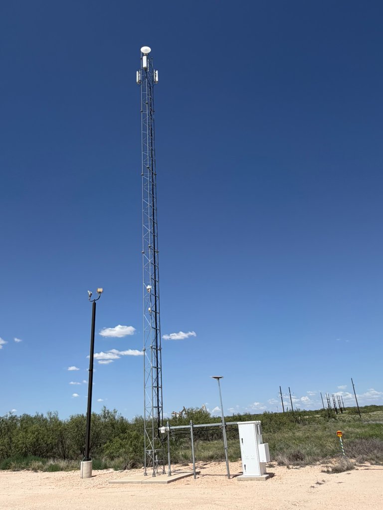 Full-height guyed lattice communication tower with equipment shelter at the base in a rural West Texas landscape