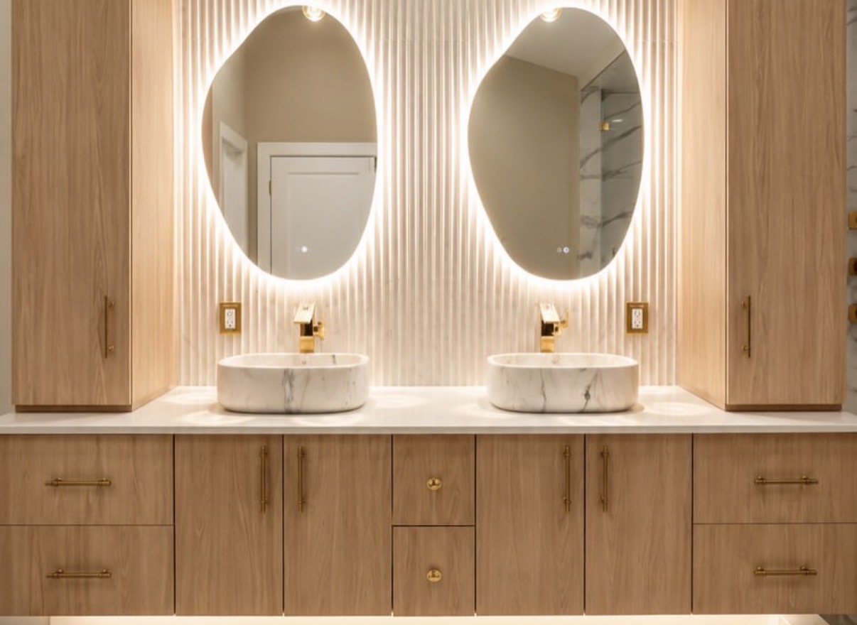 Front view of a floating double vanity with warm wood cabinetry, marble vessel sinks, brushed-gold faucets, and backlit oval mirrors