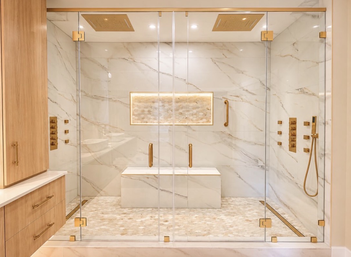 Interior of a frameless glass shower with marble-look tile, built-in bench, body sprays, and brushed-gold fixtures in this Burr Ridge, IL bathroom remodel