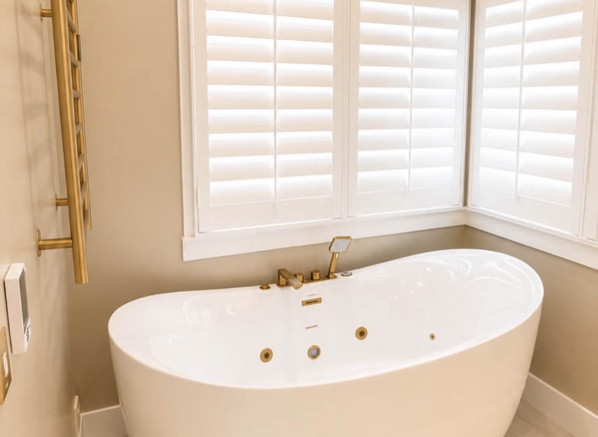 Freestanding white jetted bathtub with brushed-gold faucet, handheld sprayer, heated towel rack, and plantation shutters